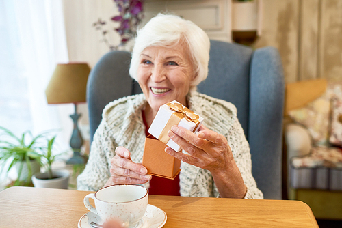 Beautiful elderly woman with charming smile sitting at wooden table and opening small gift box presented for Valentines day, stylish interior of coffeehouse on background
