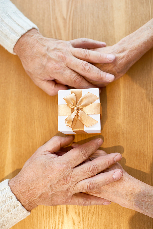 Close-up shot of loving senior couple holding hands while celebrating wedding anniversary at restaurant, small gift box lying on wooden table