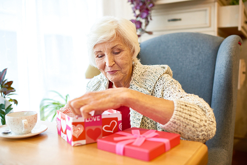 Celebrating Valentines day in restaurant: pretty senior woman wearing knitted cardigan  opening gift box with interest while sitting at wooden table with cup of fragrant coffee, portrait shot