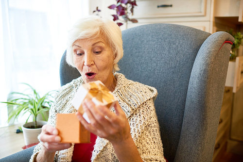 Surprised elderly woman wearing knitted cardigan unwrapping gift box while sitting on cozy armchair, portrait shot