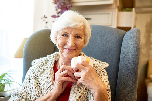 Waist-up portrait of pretty senior woman  with toothy smile while sitting on cozy armchair and holding small gift box in hands