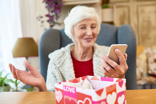 Cheerful senior woman holding modern smartphone received as Christmas present in hand while sitting on cozy armchair, interior of stylish living room on background