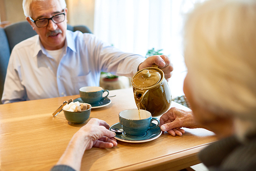 Waist-up portrait of handsome senior man pouring herbal tea in cups while having romantic date with wife, interior of cozy cafe on background