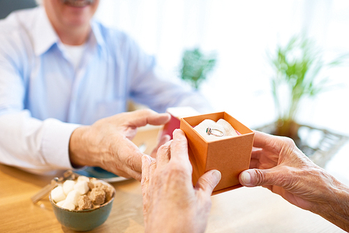Close-up shot of senior man wearing blue shirt sitting at restaurant table with gift box containing ring and making proposal to his soulmate, blurred background