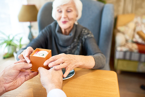 Waist-up portrait of cheerful senior woman wearing knitted sweater sitting at cafe table and enjoying beauty of ring presented as gift for their wedding anniversary, focus on foreground