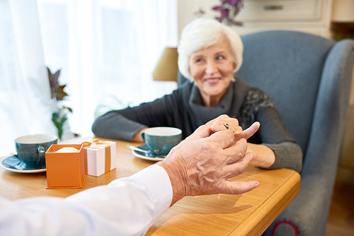 Beautiful elderly woman sitting at cafe table and looking at her fiance with toothy smile while he putting engagement ring on her finger