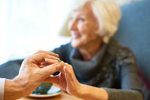 Close-up shot of elderly fiance putting engagement ring on finger of pretty woman while sitting at restaurant, focus on foreground