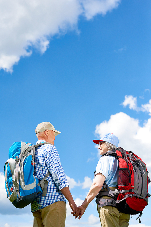 Back view portrait of loving senior couple travelling on hiking trip, both wearing backpacks, standing holding hands against clear blue sky