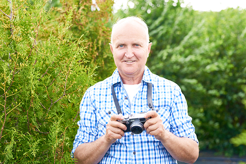 Portrait of smiling senior man taking pictures in park using vintage photo camera while travelling and 