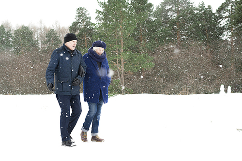 Full length  portrait of modern senior couple walking in winter forest and chatting on the way with snow falling gently, copy space