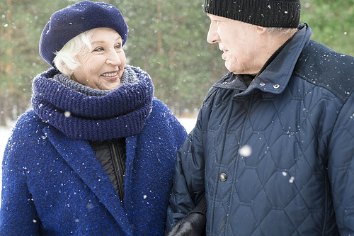 Waist up portrait of cheerful senior couple walking in winter forest and chatting happily with snow falling gently