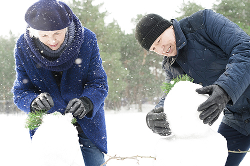 Portrait of modern senior couple building snowman while enjoying walk in beautiful winter forest with snow falling gently