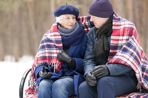 Portrait of loving senior couple sitting on bench in winter sharing blanket and chatting, copy space