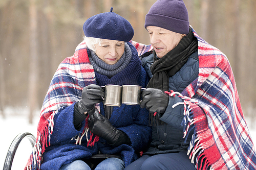 Portrait of loving senior couple drinking hot cocoa sitting on bench outdoors and sharing blanket