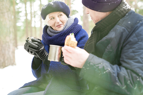 Portrait of happy senior couple enjoying picnic in snowy winter park, focus on woman smiling