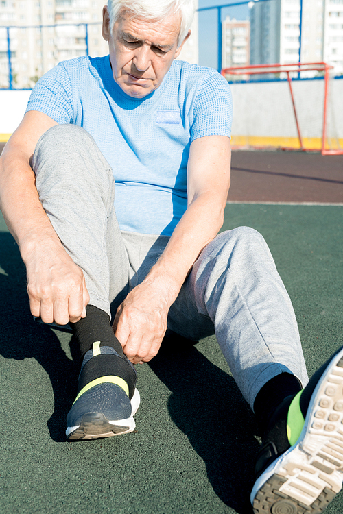Portrait of white haired senior man tying sports shoes sitting on running track outdoors