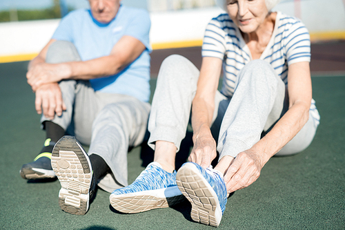 Mid section portrait of white haired senior couple tying sports shoes sitting on running track outdoors, copy space