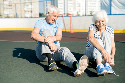 Portrait of white haired senior couple tying sports shoes sitting on running track outdoors, copy space