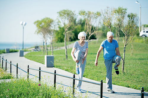 Full length portrait of active senior couple stretching legs standing on running track outdoors in sunlight looking happy, copy space
