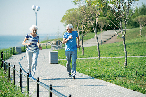 Full length portrait of active senior couple running on park lane along sea shore outdoors , copy space