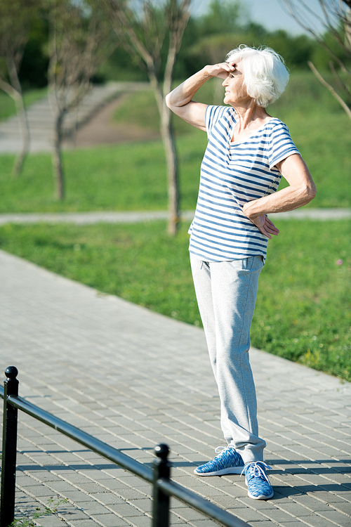 Full length portrait of active senior woman looking away shielding eyes from sunlight outdoors, copy space