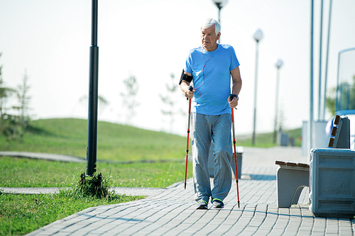 Full length portrait of active senior man practicing Nordic walking with poles outdoors in park and listening to music in earphones, copy space