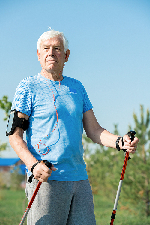 Portrait of active senior man practicing Nordic walking with poles outdoors in park and listening to music , copy space