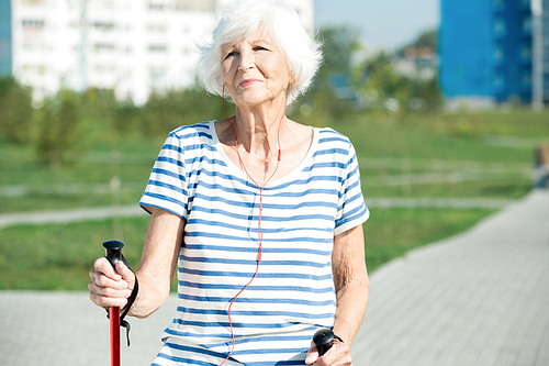 Waist up portrait of modern senior woman enjoying Nordic walking with poles in park and smiling while listening to music, copy space, sunlight,