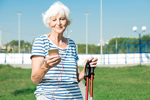 Waist up portrait of modern senior woman enjoying Nordic walking with poles in park and smiling while setting music playlist on smartphone, copy space