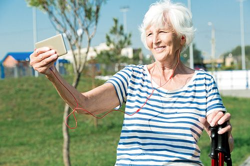 Waist up portrait of modern senior woman enjoying Nordic walking with poles in park and smiling while taking selfie, copy space