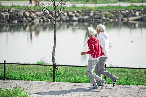 Side view full length portrait of active senior couple running in park along lake, copy space