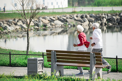 Side view portrait of active senior couple running in park along lake, copy space