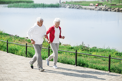 Full length portrait of senior couple running together in park along water in sunlight, copy space