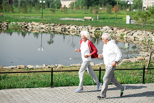 Full length portrait of senior couple running together in park along lake in sunlight, copy space