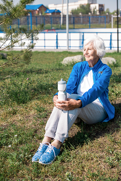 Full length portrait of modern senior woman resting sitting on lawn and smiling happily after morning run in park