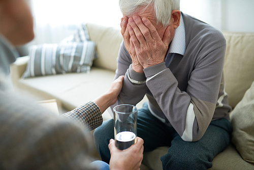 Crop compassionate person consoling elderly man in assisted living home holding glass of water.