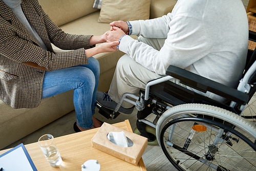 Low section of female therapist holding hands with senior patient in wheelchair, comforting him in difficult situation, copy space