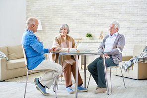 Full length portrait of three happy senior friends sitting at table enjoying tea meeting at home and sharing memories, copy space