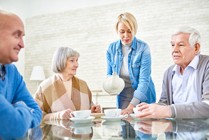 Woman pouring tea for senior patients in assisted living home sitting at table and chatting.