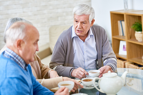 Portrait of white-haired senior man talking to friend while drinking tea at table in modern retirement home, copy space