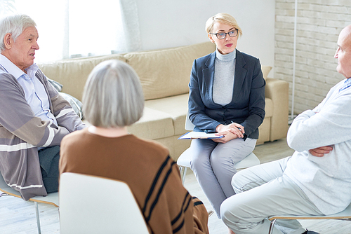 Portrait of blonde female psychiatrist wearing glasses leading group therapy session for senior people sitting in circle in retirement home, copy space