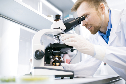 Side view portrait of smiling scientist looking in microscope while doing research in medical laboratory