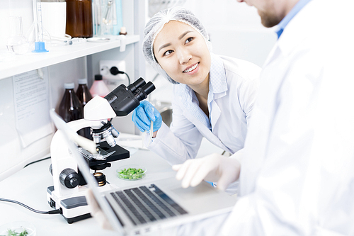 Crop view of smiling female microbiologist standing at eyepiece of microscope with samples of green vegetables looking at colleague typing results of analysis on laptop