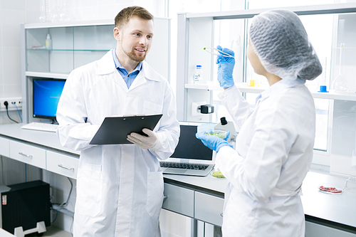 Young scientists in white laboratory coats standing at desk with laptop talking about their research, woman holding pincers with probe of tested green food fiber