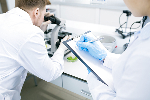 Crop back view of female microbiologist writing down into clipboard data received from man analyzing green vegetable structure under microscope