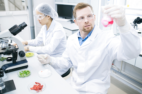 Portrait of two scientists doing research studying food substances in laboratory, focus on modern young man holding beaker