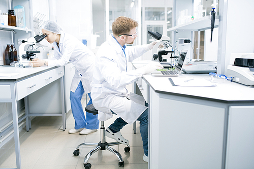 Side view of male microbiologist in glasses sitting at desk using laptop and holding hand on microscope with food samples with assistant looking at microscope eyepiece on background .