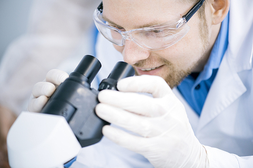 Crop portrait of smiling young scientist looking in microscope while working on research in medical laboratory