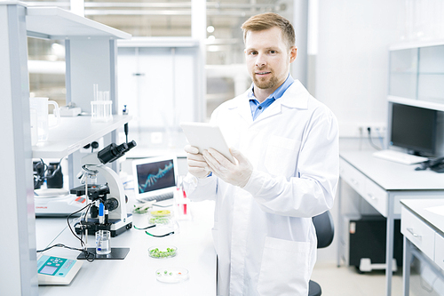 Young male microbiologist in laboratory coat standing at desk with microscope and green vegetable samples and holding tablet smiling and 