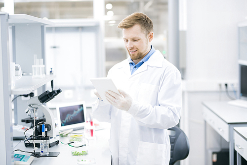 Waist up portrait of smiling young scientist wearing lab coat working on research using digital tablet, copy space